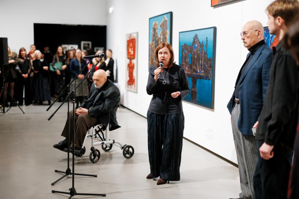 At an exhibition opening, a woman speaks into a microphone before an audience; exhibition participants stand nearby, with paintings on white walls and listeners visible in the background.