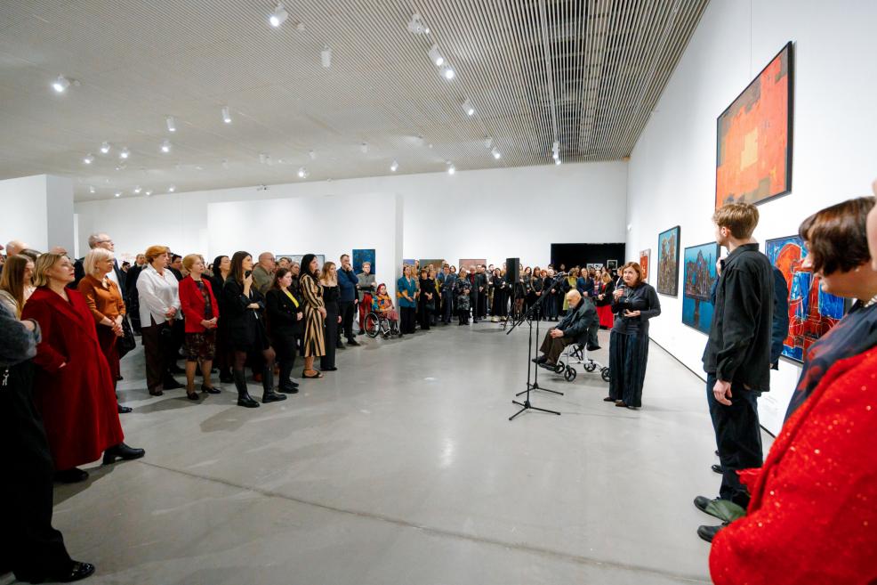 Wide view of an exhibition opening: visitors gathered in the gallery listening to a speech, microphones and a seated man in a wheelchair in the foreground, with paintings displayed on the surrounding walls.