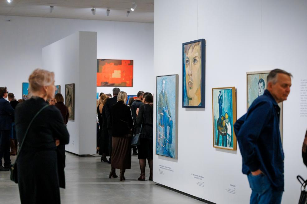 View inside the exhibition gallery with visitors moving through the space and viewing paintings on white walls; portraits and figurative works in the foreground, with a crowd visible in the background.