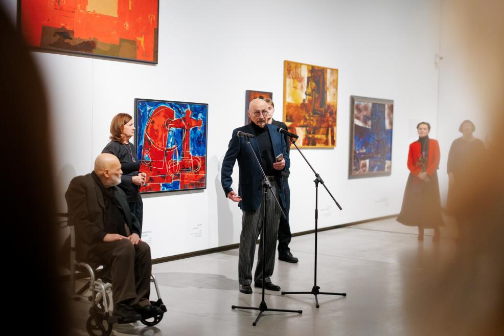 Opening moment of an exhibition in the gallery: a man speaking at a microphone to the audience, with colorful paintings on the wall behind; the artist seated in a wheelchair nearby, other participants standing alongside.