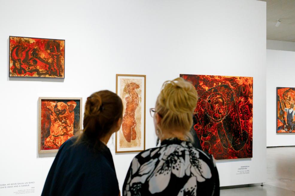 Two visitors seen from behind viewing an exhibition of paintings in red and orange tones on a white gallery wall.