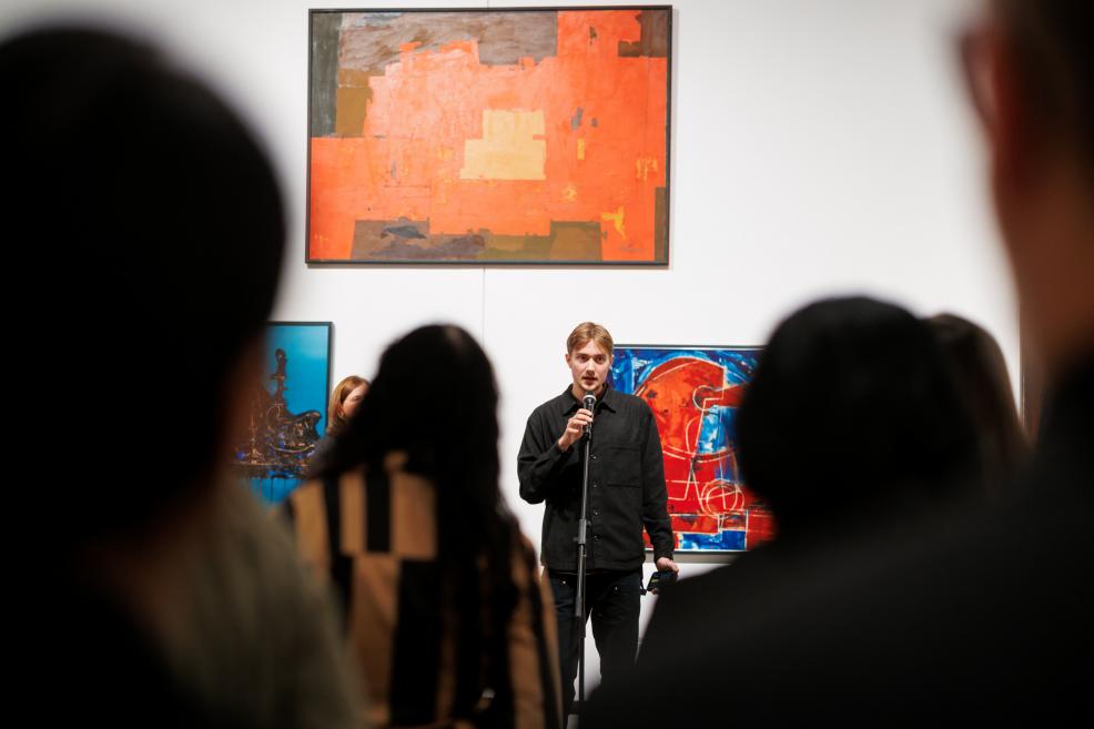 A young man speaking into a microphone at an exhibition opening, with vivid orange and blue abstract paintings in the background and the silhouettes of the audience in the foreground.