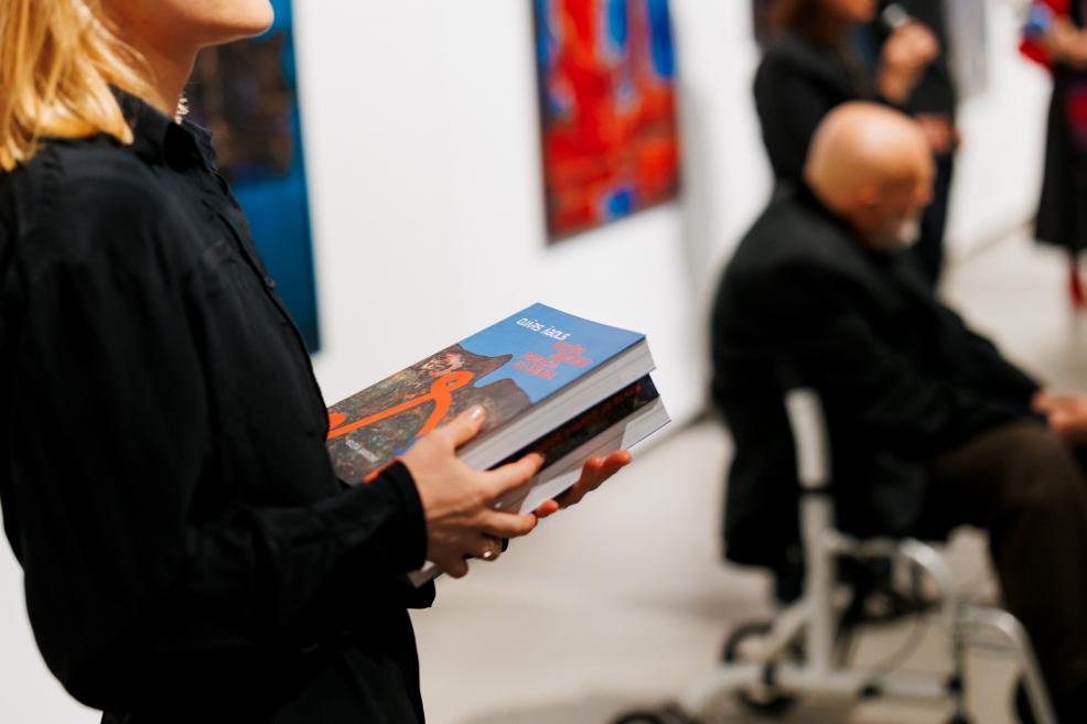 A woman dressed in black holding an exhibition catalogue featuring Ojārs Ābols’s artwork on the cover; in the background, a man seated in a wheelchair and the exhibition space with paintings.