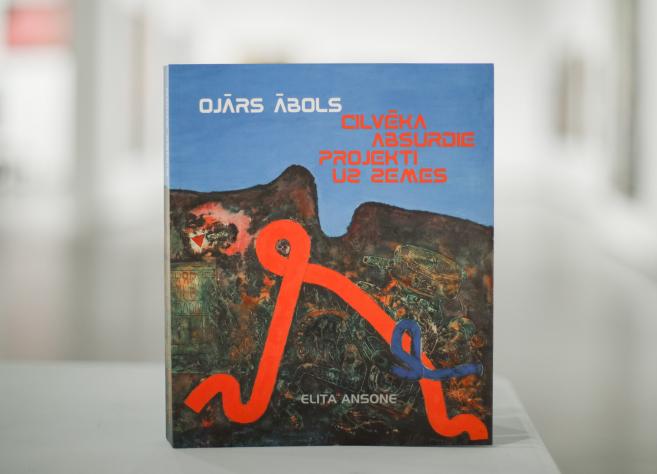 Grāmata “Ojārs Ābols. Cilvēka absurdie projekti uz zemes” ar mākslinieka darbu reprodukciju uz vāka, novietota uz galda izstāžu telpā.