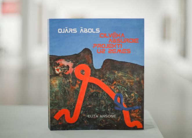Grāmata “Ojārs Ābols. Cilvēka absurdie projekti uz zemes” ar mākslinieka darbu reprodukciju uz vāka, novietota uz galda izstāžu telpā.