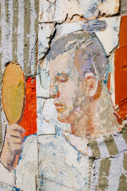 Fragment of a wall painting showing a stylized male profile holding a round mirror, with peeling paint layers and visible cracks.