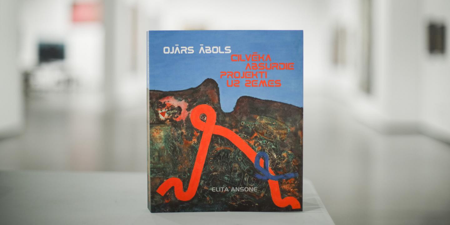 Grāmata “Ojārs Ābols. Cilvēka absurdie projekti uz zemes” ar mākslinieka darbu reprodukciju uz vāka, novietota uz galda izstāžu telpā.