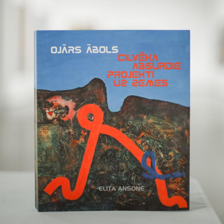 Grāmata “Ojārs Ābols. Cilvēka absurdie projekti uz zemes” ar mākslinieka darbu reprodukciju uz vāka, novietota uz galda izstāžu telpā.