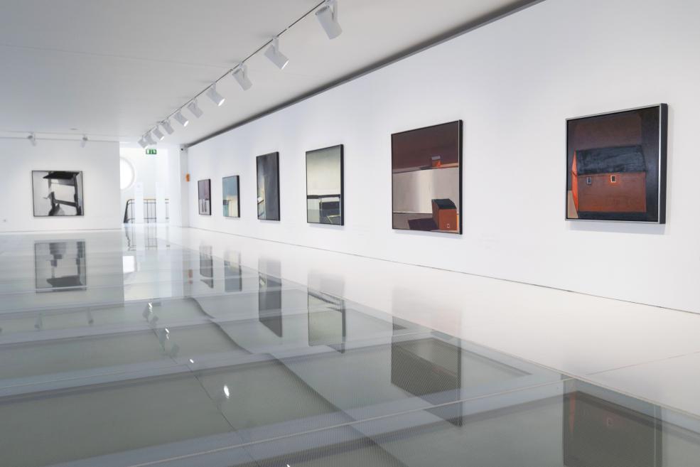 Mūsdienu mākslas galerijas telpa ar baltām sienām, pie sienas izkārtotām gleznām un spīdīgu grīdu, kurā atspīd ekspozīcija.