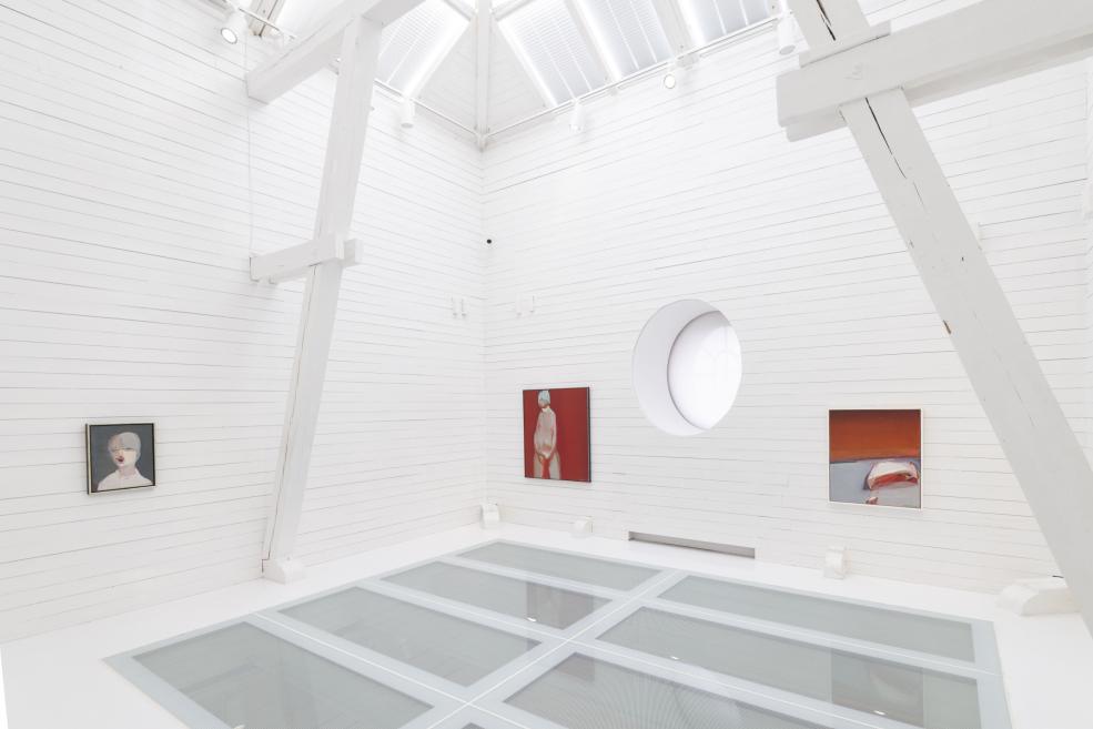 Balta galerijas telpa ar trim sienas gleznām, lielām baltām sijām un apaļu logu augstajā sienā.