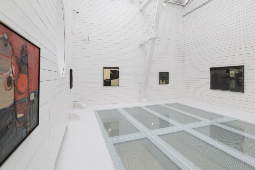 Balta galerijas telpa ar vairākām abstraktām gleznām uz sienām un stiklotu grīdas laukumu.