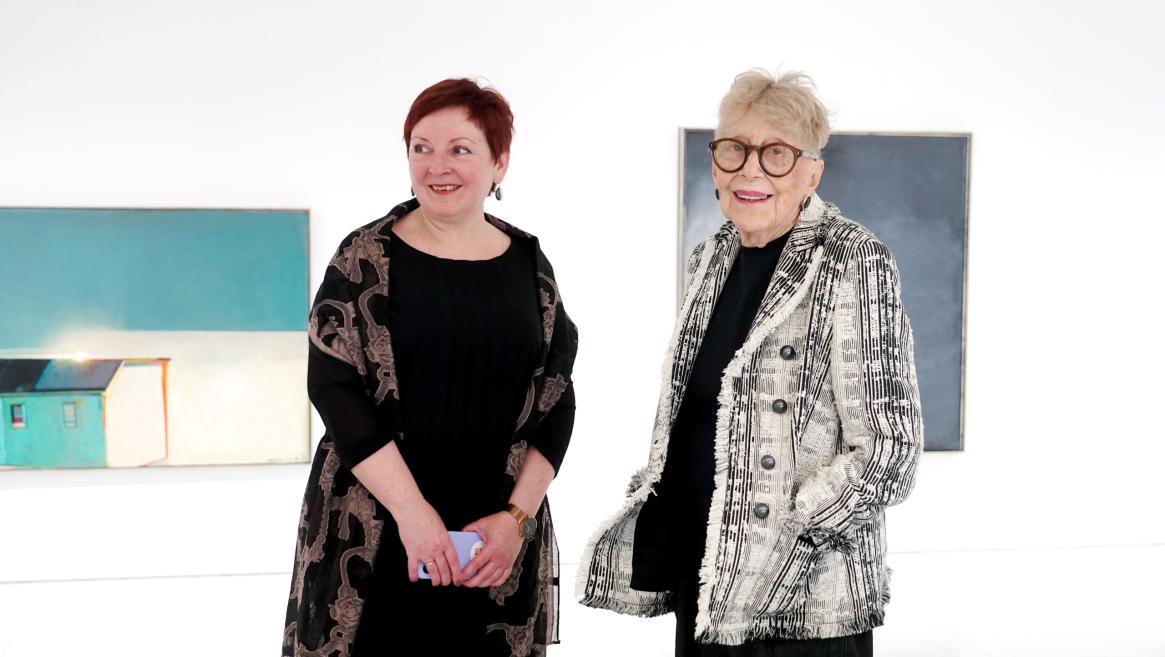 Divas sievietes stāv pie sienas baltā mākslas galerijā ar gleznām fonā.