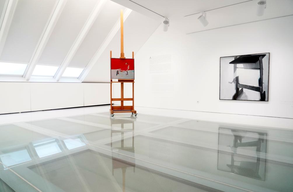 Gaišā mākslas galerijas telpā ar slīpu baltu griestu redzams molberts ar gleznu un pie sienas piekārts melnbalts darbs, kas atspoguļojas spīdīgajā grīdā.