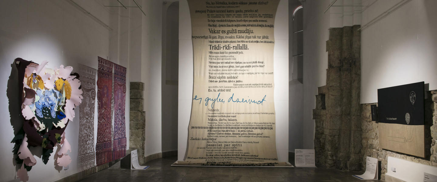 In an exhibition space with historic stone walls and vaulted architecture, a large suspended text installation hangs at the center, surrounded by contemporary artworks including textiles, wall-mounted pieces, and graphic works.