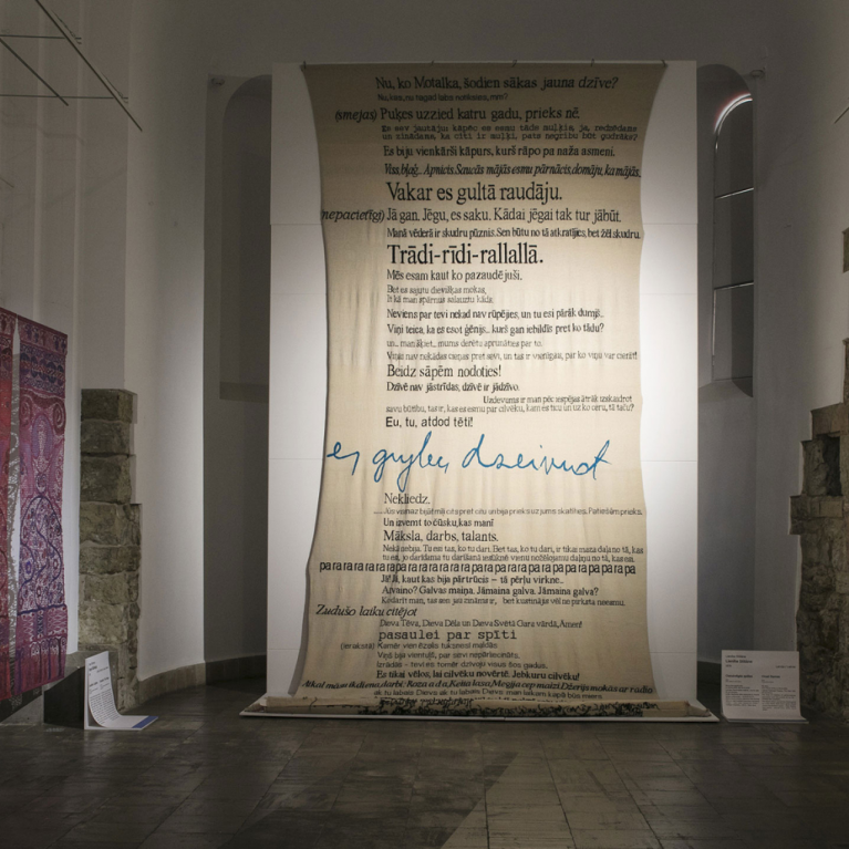 In an exhibition space with historic stone walls and vaulted architecture, a large suspended text installation hangs at the center, surrounded by contemporary artworks including textiles, wall-mounted pieces, and graphic works.