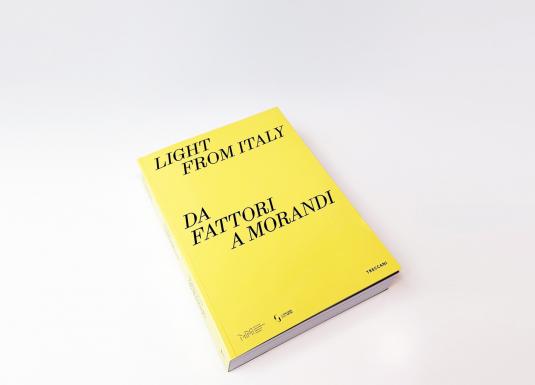 Uz gaišas virsmas novietots dzeltens izstādes katalogs ar uzrakstu “Light from Italy. Da Fattori a Morandi”.
