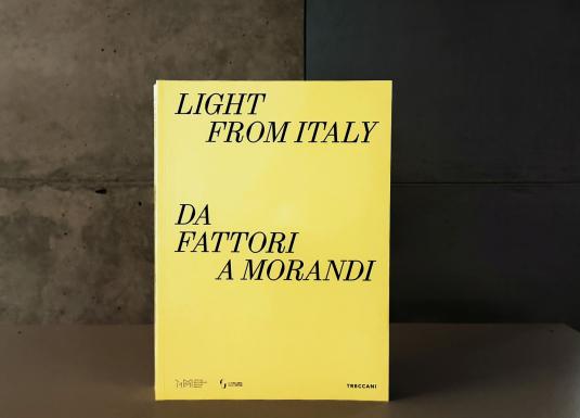 Dzeltenas krāsas grāmatas vāks ar melnu uzrakstu “Light from Italy. Da Fattori a Morandi”, novietots uz galda mūsdienīgā muzeja telpā; fonā redzama betona siena un kāpnes.
