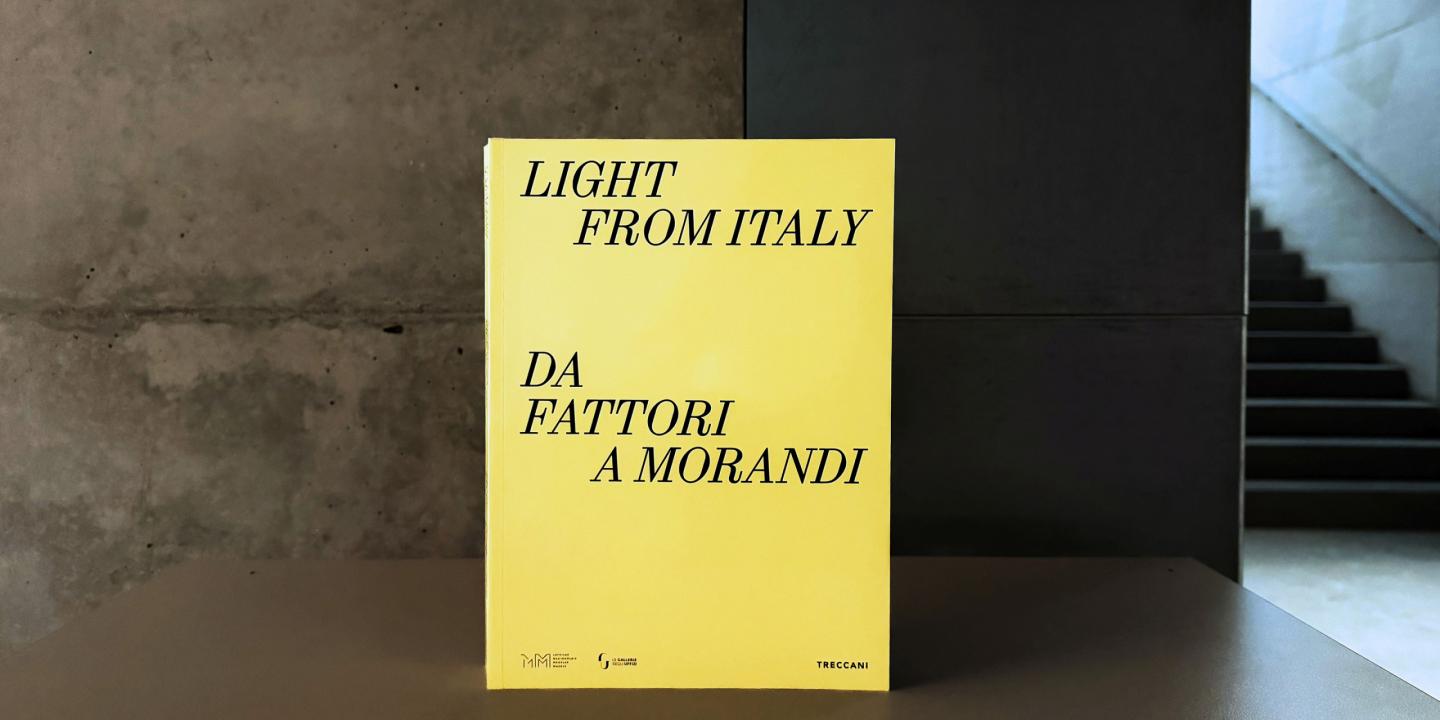 Dzeltenas krāsas grāmatas vāks ar melnu uzrakstu “Light from Italy. Da Fattori a Morandi”, novietots uz galda mūsdienīgā muzeja telpā; fonā redzama betona siena un kāpnes.
