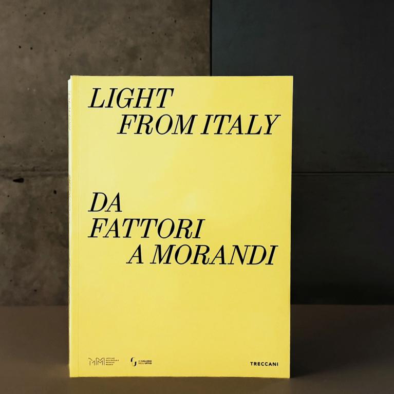 Dzeltenas krāsas grāmatas vāks ar melnu uzrakstu “Light from Italy. Da Fattori a Morandi”, novietots uz galda mūsdienīgā muzeja telpā; fonā redzama betona siena un kāpnes.