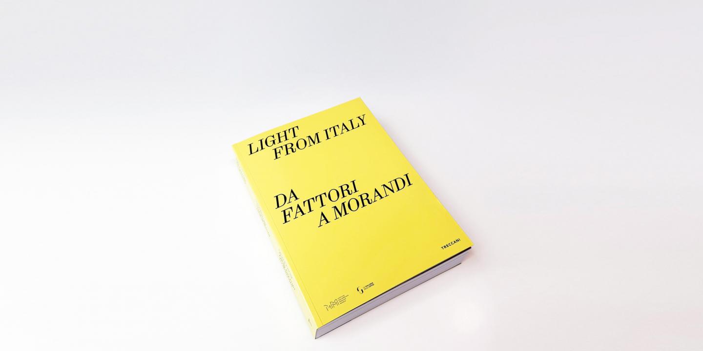 Uz gaišas virsmas novietots dzeltens izstādes katalogs ar uzrakstu “Light from Italy. Da Fattori a Morandi”.