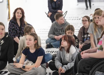 Muzeja zālē uz grīdas sēž bērnu grupa un divas pieaugušas sievietes, tostarp viena ratiņkrēslā, klausoties nodarbībā vai sarunā.