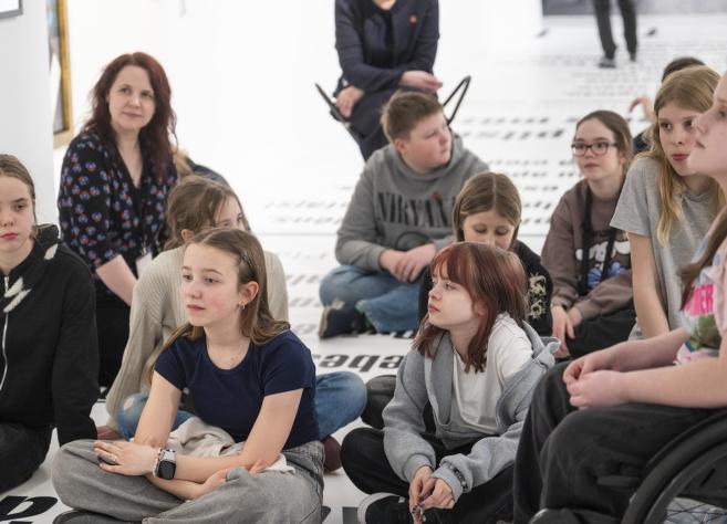 Muzeja zālē uz grīdas sēž bērnu grupa un divas pieaugušas sievietes, tostarp viena ratiņkrēslā, klausoties nodarbībā vai sarunā.