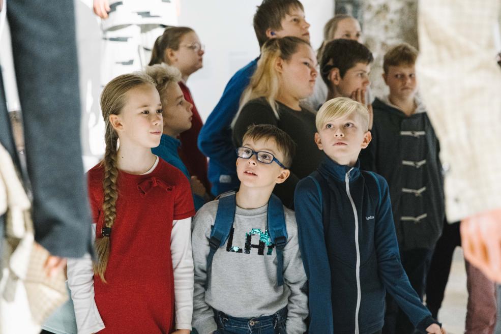 Muzeja zālē stāv bērnu grupa un skatās uz kaut ko ārpus kadra.