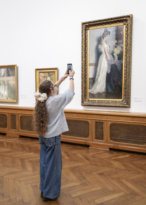 Meitene ar garām bizēm un lielu matu banti muzeja zālē fotografē ar telefonu lielu gleznu zeltītā rāmī.
