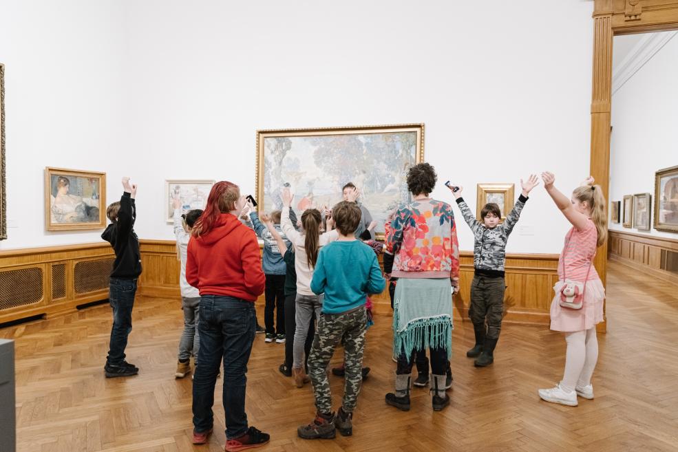 Bērnu grupa ar paceltām rokām stāv mākslas muzeja zālē pie sienām izvietotām gleznu un darbu rāmjos.
