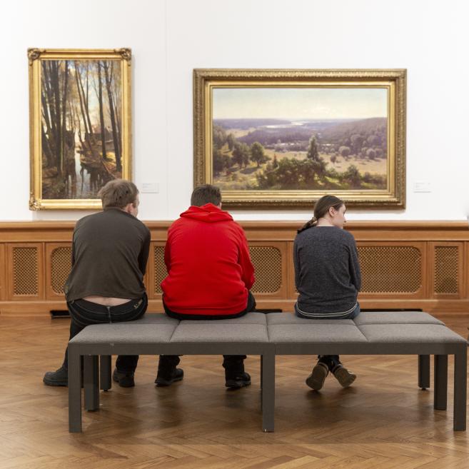 Trīs cilvēki sēž uz sola mākslas galerijā pretī divām ierāmētām ainavu gleznām uz baltas sienas.