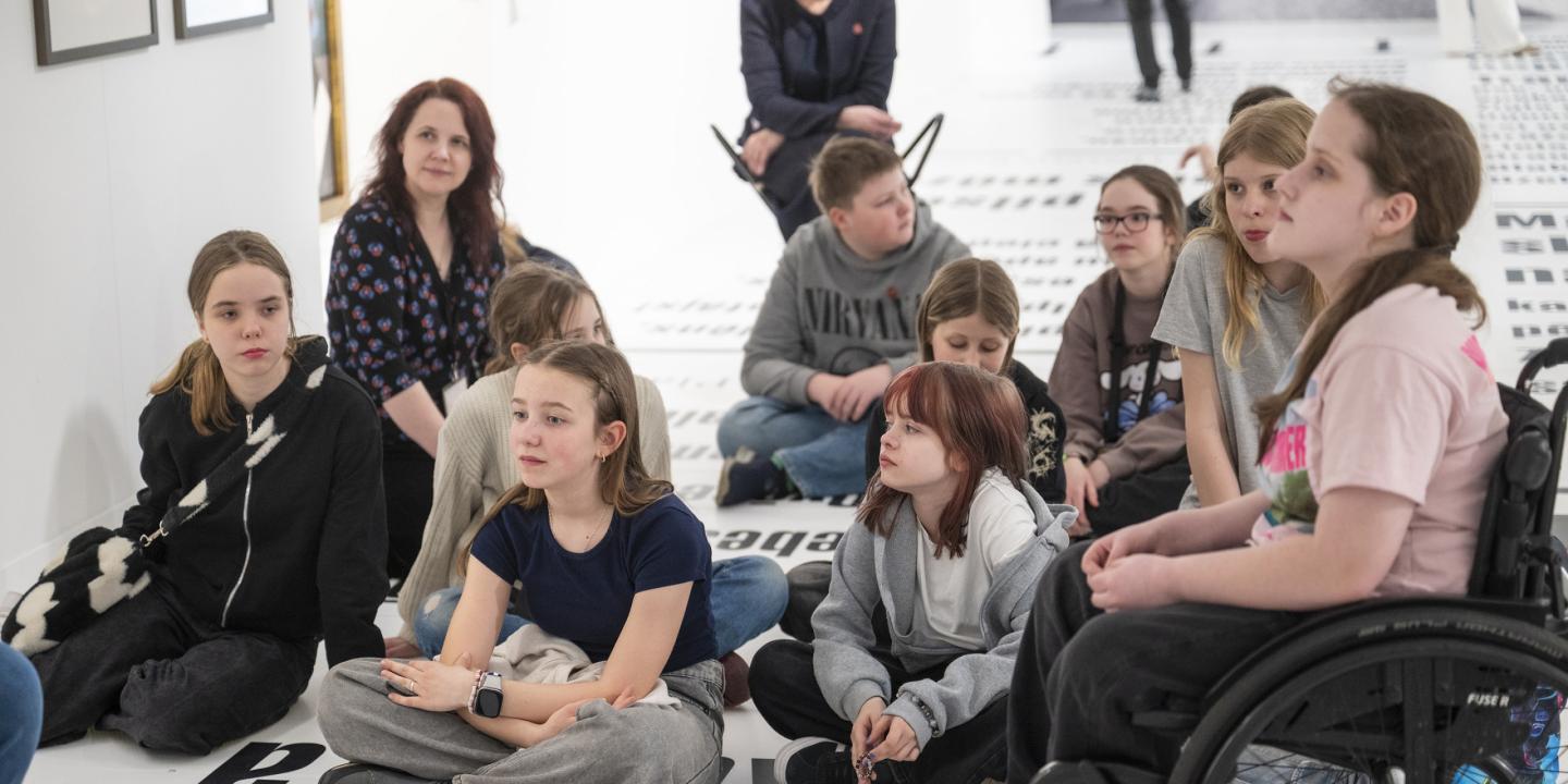 Muzeja zālē uz grīdas sēž bērnu grupa un divas pieaugušas sievietes, tostarp viena ratiņkrēslā, klausoties nodarbībā vai sarunā.