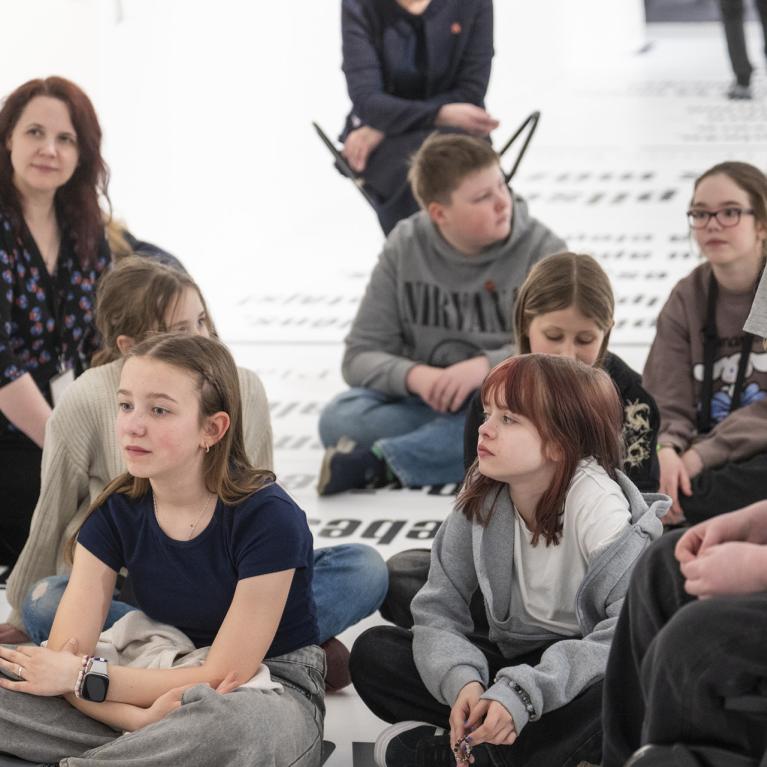 Muzeja zālē uz grīdas sēž bērnu grupa un divas pieaugušas sievietes, tostarp viena ratiņkrēslā, klausoties nodarbībā vai sarunā.
