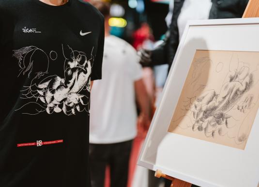 Mākslas darbs ar zīmējumu, kuram blakus ir krekls, kurā attēlots šis zīmējums