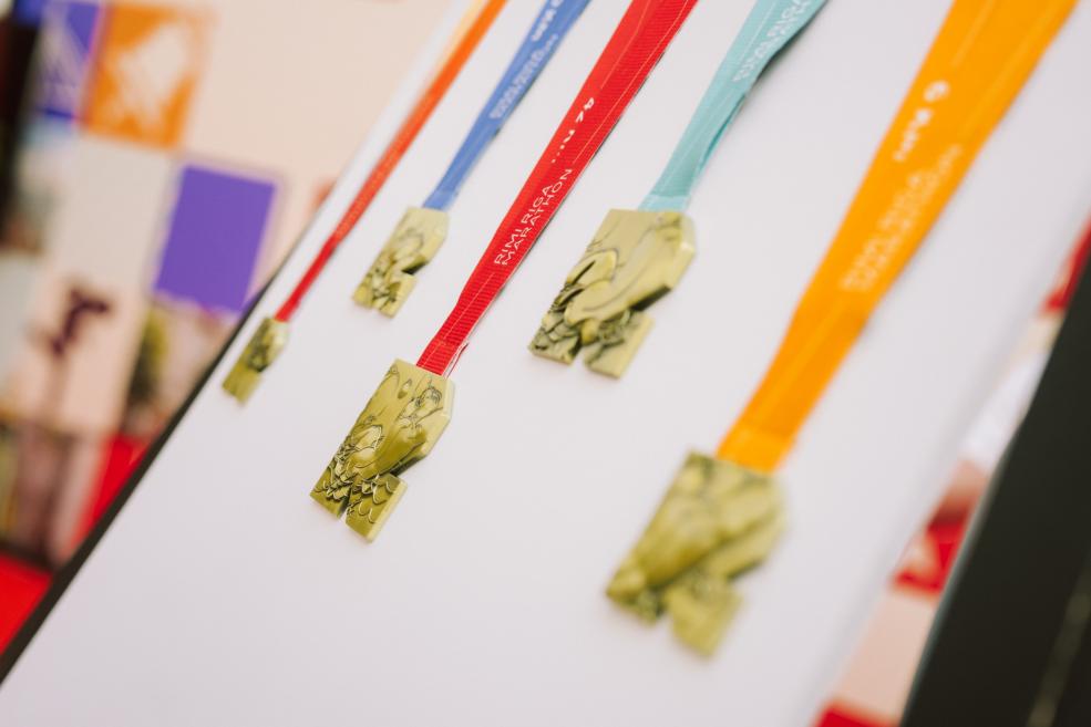 Vairākas zelta krāsas medaļas ar dažādu krāsu lentēm izkārtotas uz balta stenda.