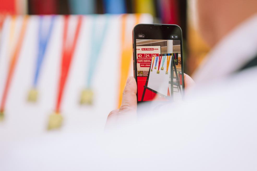 Persona fotografē medaļas ar krāsainām lentēm, izmantojot viedtālruni.