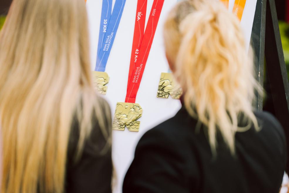 Divas personas no mugurpuses aplūko vairākas medaļas ar krāsainām lentēm, izkārtotas uz stenda.