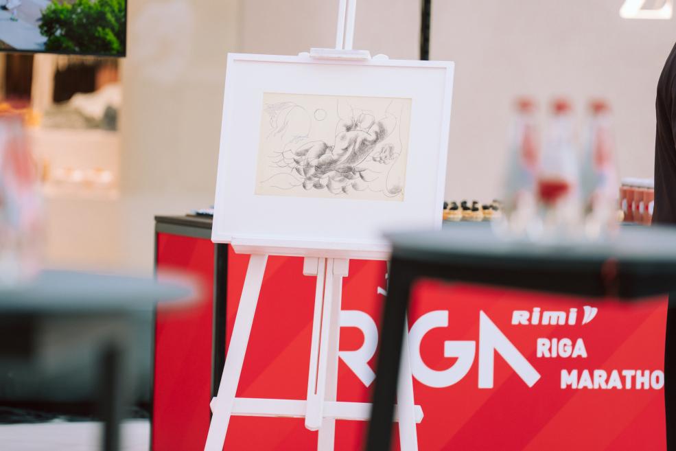 Uz statīva izstādīts melnbalts zīmējums, fonā redzams “Rimi Rīga Marathon” stends.