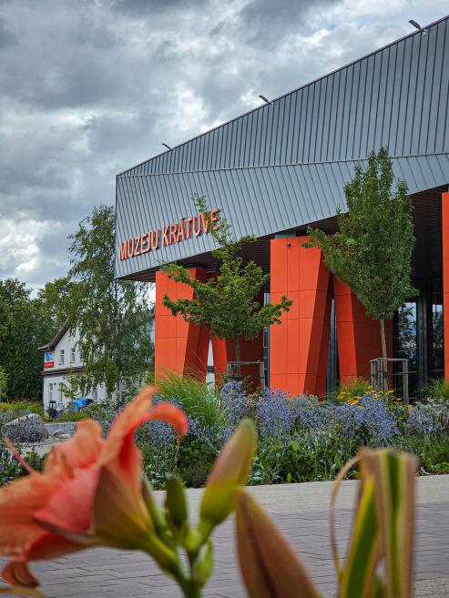 Mūsdienīga muzeju krātuves ēka ar oranžiem balstiem un uzrakstu “Muzeju krātuve”, priekšplānā ziedi.