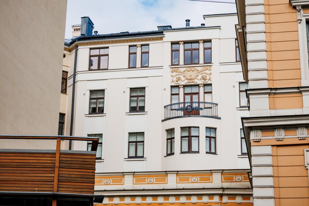 Skats uz vēsturiskas ēkas fasādi ar dekoratīviem elementiem, logiem un nelielu balkonu pilsētvidē.