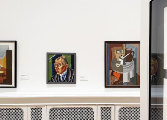 Muzeja zāles sienā izvietotas vairākas gleznas, tostarp portrets un klusā daba, baltā interjerā.