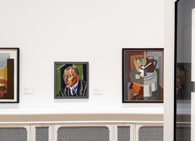 Muzeja zāles sienā izvietotas vairākas gleznas, tostarp portrets un klusā daba, baltā interjerā.