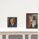 Muzeja zāles sienā izvietotas vairākas gleznas, tostarp portrets un klusā daba, baltā interjerā.