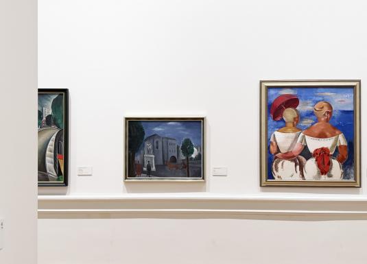 Muzeja sienā izkārtotas vairākas gleznas, tostarp ainava un divu figūru kompozīcija spilgtās krāsās.