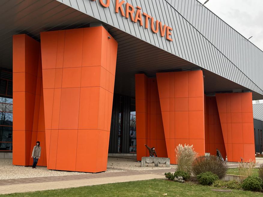 Muzeju krātuves ēkas fasāde ar lieliem oranžiem balstiem un uzrakstu “Muzeju krātuve”.