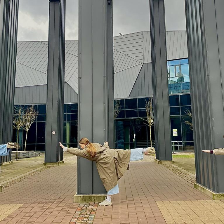 Trīs jaunieši pie muzeja ēkas kolonnām izpilda līdzsvara pozu ar izstieptām kājām un rokām.