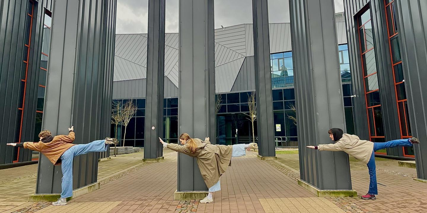 Trīs jaunieši pie muzeja ēkas kolonnām izpilda līdzsvara pozu ar izstieptām kājām un rokām.