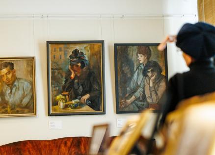 Muzeja sienā izkārtotas trīs gleznas ar cilvēku portretiem, kamēr priekšplānā apmeklētāja žestikulē, skatoties uz darbiem.