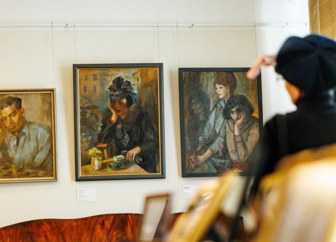 Muzeja sienā izkārtotas trīs gleznas ar cilvēku portretiem, kamēr priekšplānā apmeklētāja žestikulē, skatoties uz darbiem.