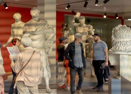 Muzeja zālē ar klasiskām skulptūrām apmeklētāji aplūko eksponātus un lasa informācijas materiālus.