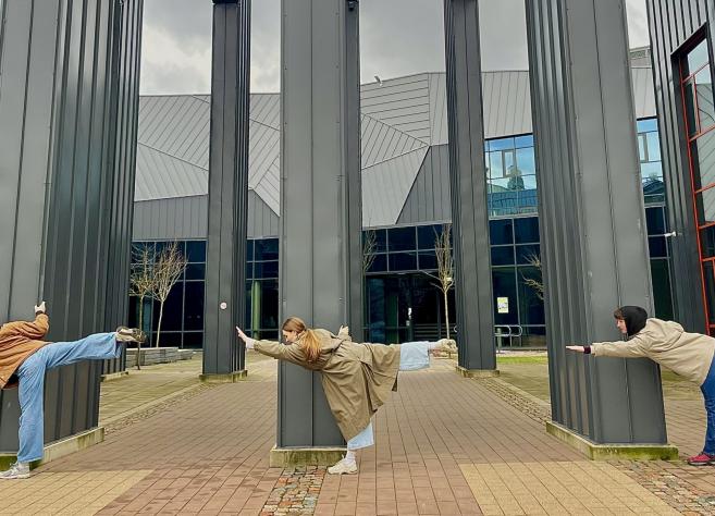 Trīs jaunieši pie muzeja ēkas kolonnām izpilda līdzsvara pozu ar izstieptām kājām un rokām.